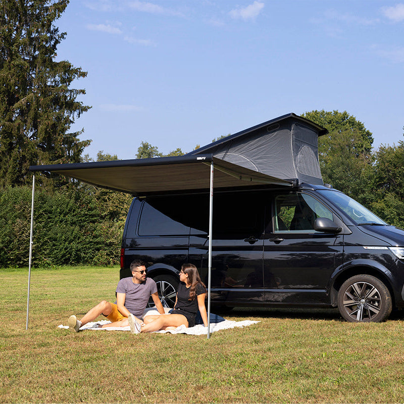Two people sitting on a blanket in front of a black camper van with an extended Fiamma F43 awning in a grassy area with trees.