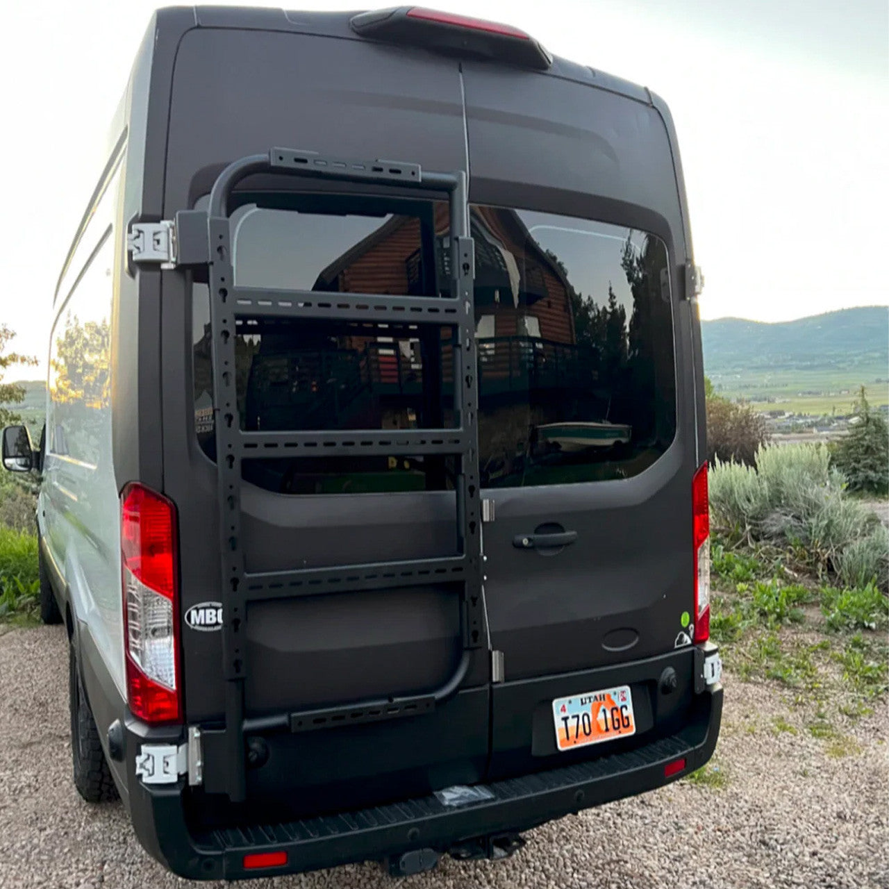 Back view of a black van with a ladder on a gravel road with a scenic background